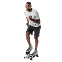 A man in a light gray t-shirt and dark gray shorts is running on a stair stepper machine against a solid white background.