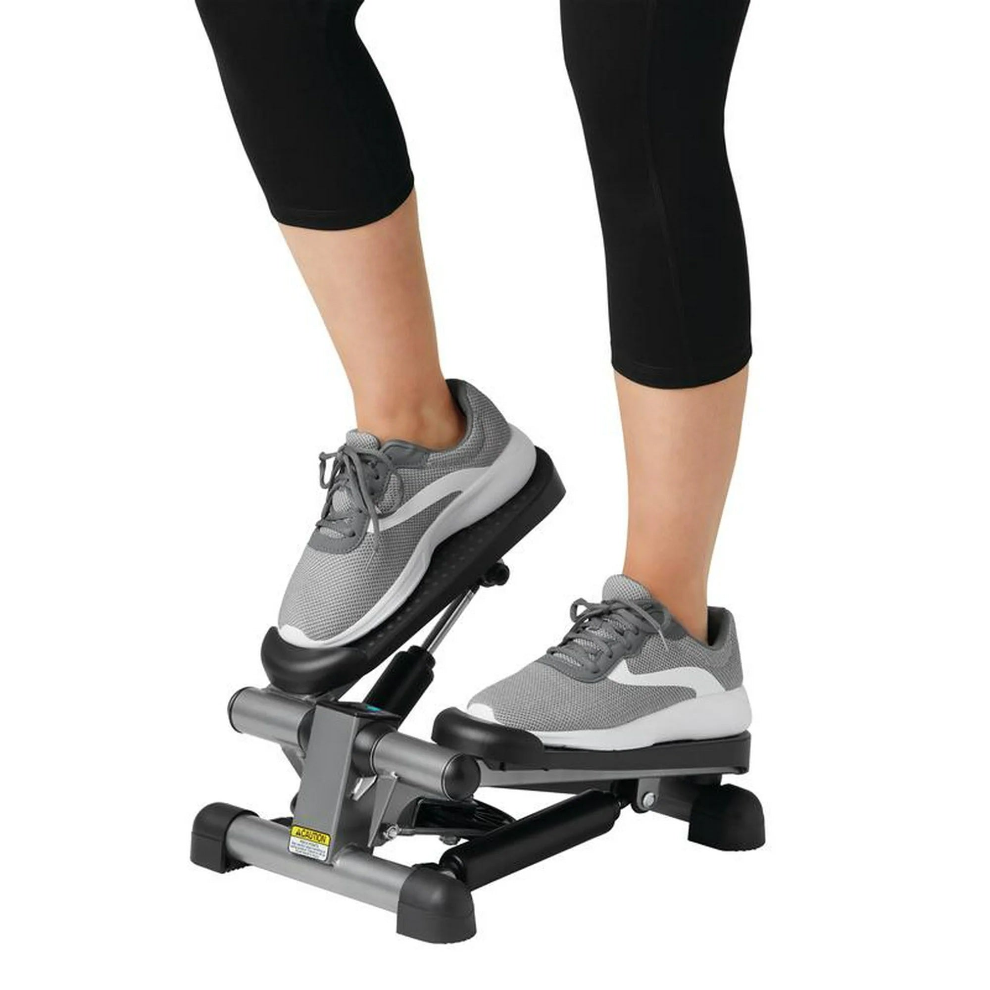 A person using a stair stepper machine, wearing gray and white athletic sneakers and black capri leggings, against a plain white background.