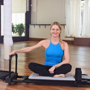 Middle age woman seated on pilates while holding the right hand on footbar.