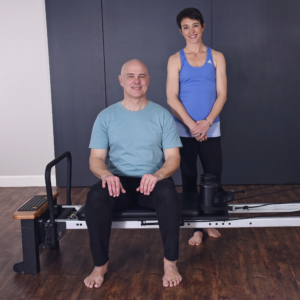 Middle age man sitting on padded seat and woman standing behind the pilates.