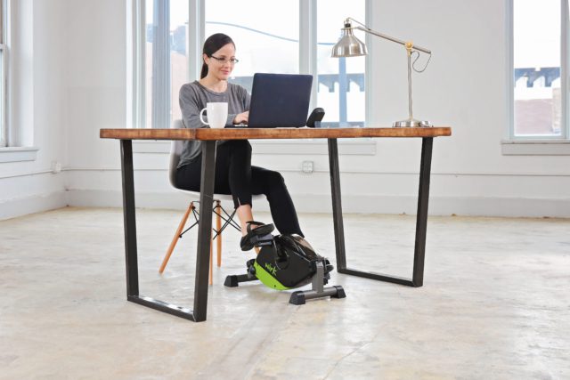 A woman uses a compact exercise bike under her desk at home.